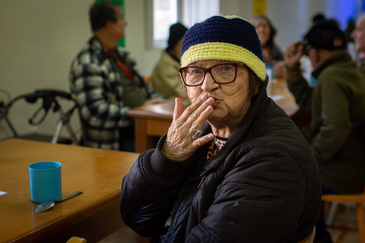 Moradora de óculos e gorro enviando um beijo no refeitório do Asilo.