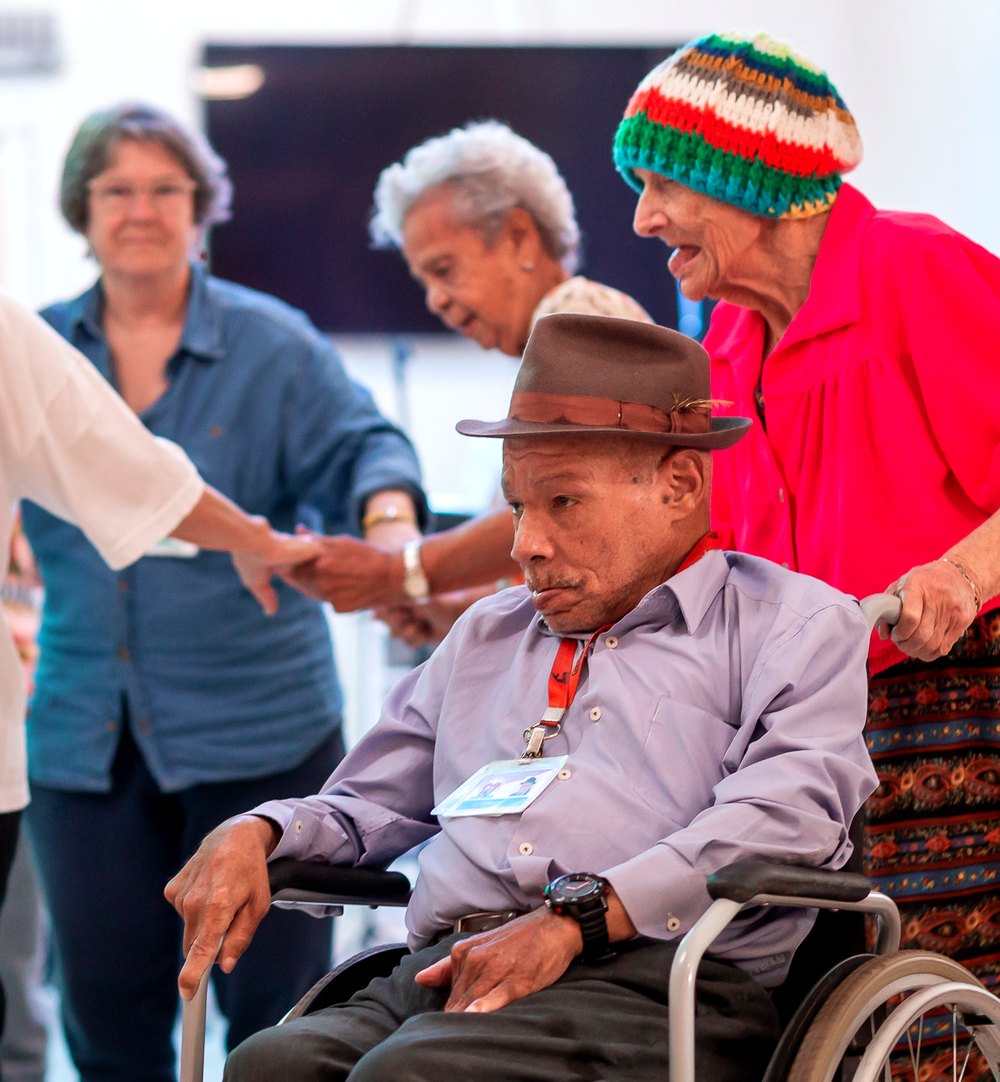 Moradores do Asilo Padre Cacique dançando juntos. À frente, um morador em cadeira de rodas conduzido por uma moradora.